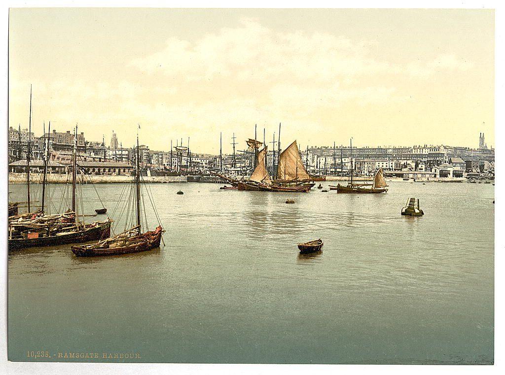 The image depicts a historic view of Ramsgate Harbour in the early 19th century. It shows various sailing vessels, including large ships with tall masts and smaller boats, moored within calm waters near the harbor's edge. The background features an urban landscape with numerous buildings indicative of industrial or commercial establishments typical for that era. Notable is a prominent sailboat with its sails unfurled in full against the partly cloudy sky. Various buoys are floating on the water to mark channels and depths, hinting at navigational aids used during this time period. The overall composition suggests an active port bustling with maritime activity amidst urban growth.