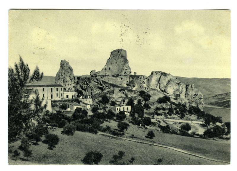 The image shows a monochrome photograph of an ancient landscape with large rock formations and what appears to be ruins or old structures perched atop them. The foreground includes scattered trees, shrubs, and some buildings that seem like historical estates or farmhouses. The middle ground features more extensive rocky outcrops leading up to the elevated areas where the prominent rocks stand. In the background, a vast expanse of rolling hills stretches into the distance under an overcast sky. This scene likely represents a historical site with significant geological and architectural interest, possibly captured during a time when photography was not yet widely accessible or popularized as it is today.