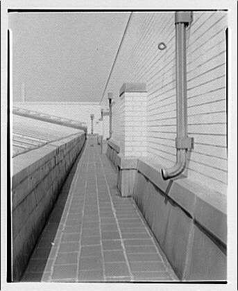 The image depicts a black and white photograph of an outdoor corridor with brick flooring, flanked by tall structures that appear to be part of a larger building. The architecture includes vertical lines and rectangular shapes, creating a sense of geometric precision.

On the left side, there's a high wall topped with metal railings or fencing for safety purposes. Attached to this structure is what looks like an overhanging roof supported by several pillars at regular intervals along the corridor length. These supports are connected vertically through beams that add stability and provide architectural interest.

The right-hand side features another elevated section, possibly part of a different building adjoining the first one or perhaps even extending from it if seen in perspective. This extension has its own set of vertical lines created by rows of windows or openings running parallel to each other at consistent intervals.

A distinctive feature is an L-shaped downspout pipe that runs along this side structure, hinting towards possible water drainage purposes. The sky above seems overcast with no visible sunlit patches; however, the overall brightness suggests daylight conditions without direct sunlight exposure on any part of the scene captured within this photograph.

The image's monochromatic color scheme and clarity offer a timeless quality to it while also emphasiz [...]