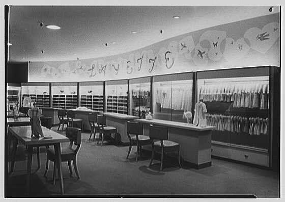 The image depicts a vintage photo of an interior space, likely from the 1940s era. The setting appears to be a department store's layette department as indicated by the caption which mentions "Layette department" along with additional context such as business locations and dates. In this specific photograph, we see rows of display cases housing various clothing items, possibly baby clothes or linens given their delicate appearance.

The layout is spacious with multiple counters running alongside glass-enclosed displays that stretch from the foreground into the background. The store's lighting seems bright, illuminating both merchandise and a few mannequins placed strategically within the space for visual merchandising purposes.

The photo is in black-and-white, which enhances its vintage feel. There are no visible customers or staff, suggesting either an off-peak hour of day when business was not bustling or that they were out of frame. The absence of any modern-day technology like digital displays hints at a time before the digital age dominated retail.

A notable feature is the overhead signage with what appears to be decorative text and possibly design elements such as hearts, which likely serve both an aesthetic purpose and are part of branding for this particular department or store chain.

The overall impression from this image captures a slice-of-life moment in mid [...]