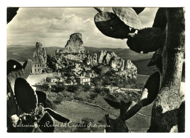 The image depicts a black and white photograph showcasing the dramatic landscape of Caltanissetta, with particular emphasis on Ruderi del Castello Pietrarossa. In the foreground, parts of cacti branches are visible, adding texture to the composition.

Dominating the midground is an imposing rocky outcrop featuring jagged formations that tower above a cluster of rustic buildings and greenery spread across undulating terrain. The architecture includes small structures with flat roofs which suggests historical or rural significance in this setting.

The background reveals a more expansive view of the region, indicating it's part of larger mountainous range under an overcast sky. There is no visible human presence in the scene, emphasizing its serene and untouched quality.

A caption on the image reads "Caltanissetta - Ruderi del Castello Pietrarossa," providing context for the location captured within this historical photograph.
