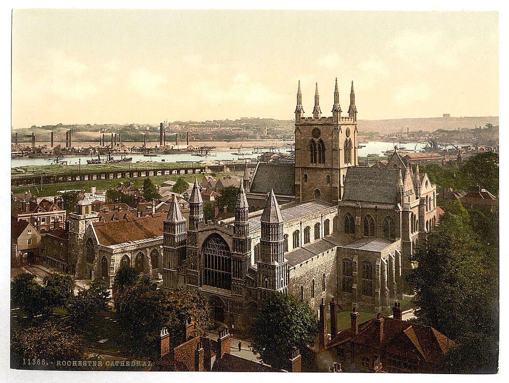 The image depicts a historical view of Rochester Cathedral in England. This Gothic-style cathedral stands prominently at the center, with its detailed architecture featuring pointed arches and intricate stonework. In the background is an expansive river with various boats and docks along the waterfront, indicating industrial activity typical for that era. Surrounding buildings exhibit varying styles from Tudor to Victorian, suggesting a town rich in history and development over centuries. The coloration of sepia tones lends authenticity and age to this image, capturing it as likely taken during late 19th or early 20th century photography processes.