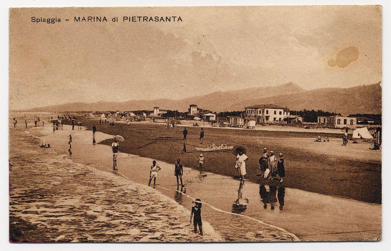 The image shows a sepia-toned photograph of what appears to be an old postcard depicting a beach scene. The label at the top reads "Spiaggia - MARINA di PIETRASANTA," which suggests that this place is known as Pietrasanta Beach or Marina in the town called Pietrasanta, possibly located on the coast.

The landscape includes several people engaged in various activities typical of a seaside setting: walking along the shoreline, sitting near water's edge, and standing with parasols. The beach stretches into the distance towards buildings that likely represent establishments such as hotels or cafes common to coastal towns during this era. There is also a hint of mountains on the horizon which might suggest the presence of an elevated geographical feature nearby.

The image appears aged, possibly dating back several decades if not more. It offers a glimpse into historical beach life and architecture from Pietrasanta's past. This photograph could be valuable for understanding the social practices and leisure activities associated with seaside towns during its time period.

For further information on this topic or similar subjects in Pieterasanta-Capezzano, including images of Ferruccio Candilera at his full work location as referenced by "https://images.loener.nl/FerroCandilera/full/6731/6731b6f689b10e7a26c6b7a9.jpg," please refer to the provided URL for additional context and d [...]