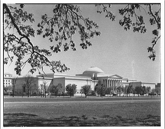 The image shows a black and white photograph of the National Gallery building, taken from an angle that captures both its front facade and part of the surrounding landscape. The gallery is a large, neoclassical structure with prominent features such as columns at the entrance and what appears to be a dome-like roof on one section. There are trees in the foreground partially obscuring some branches but allowing for clear visibility through them towards the building. A wide open space or field can be seen stretching out before the gallery, indicating that it is located within an expansive area with ample room around its perimeter. The sky above is overcast and without visible clouds, suggesting a cloudy day at the time of photography.