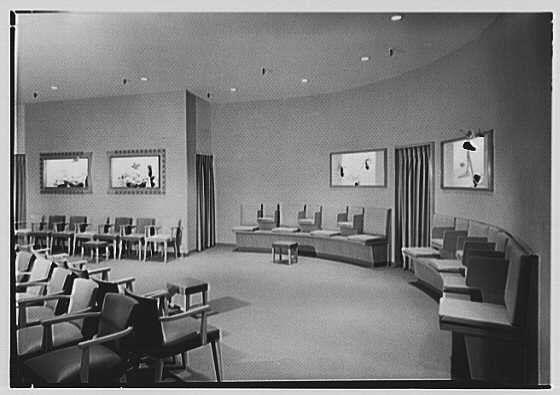 The image is a black and white photograph of an interior space, likely from the early 20th century based on its style. The room appears to be designed for exhibitions or displays, with several pieces of modernist artwork mounted on the walls above long tables. These artworks feature abstract shapes and forms, possibly intended as sculptures or relief panels.

The furniture consists of rows of chairs arranged in a semi-circle around each table, suggesting an interactive display where visitors can walk up to examine the art closely. The flooring is carpeted, which adds a sense of warmth to the room's atmosphere. Recessed lighting from ceiling fixtures provides illumination for both the artwork and seating areas.

The overall design conveys a minimalist aesthetic with clean lines and functional furniture that emphasizes viewing comfort without distracting decoration. The image lacks people or any identifying marks indicating location or specific event, keeping its focus on the layout of space itself rather than human interaction within it.