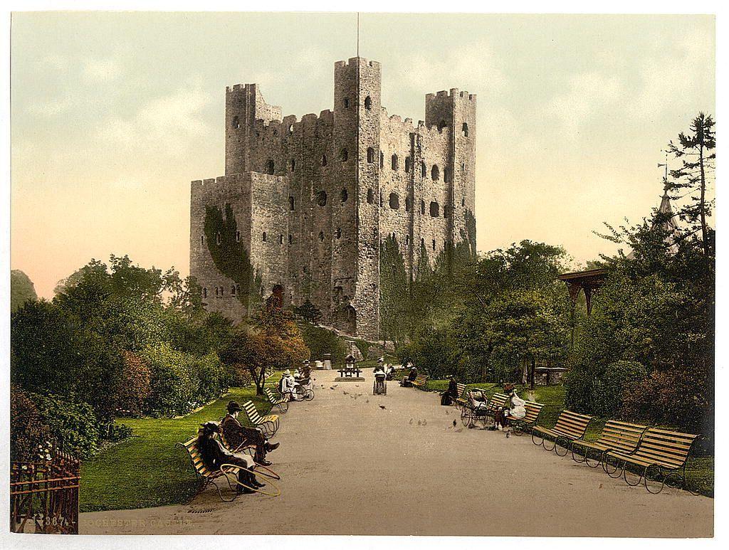 The image depicts a historical castle set amidst lush greenery, with several people leisurely strolling or seated on benches in what appears to be an old park. The architecture of the castle is characterized by its sturdy stone walls and towers, indicative of medieval design intended for defense as well as grandeur. The scene evokes a sense of tranquility and historical charm.

The coloration suggests that it's either early morning or late afternoon due to the soft light casting gentle shadows on the surroundings. People are dressed in attire reminiscent of the 19th century, hinting at the time period when this photo was likely taken. The presence of wooden benches with wrought iron detailing and decorative fencing adds a touch of elegance to the park setting.

This particular image is part of a collection titled "Views of the British Isles England Kent Rochester," which indicates that it's one among many capturing various aspects of English heritage, specifically focusing on Kent in East Sussex, where Rochester Castle resides. The reference number '65e4' might be an internal cataloging system or identification code for this specific image within a larger collection.

Further details such as the exact date and context behind these photos would require additional research beyond what is visible in the provided image caption from "The castle, Rochester, England between ca.  [...]