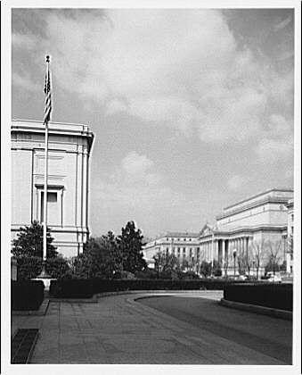 The image depicts a black and white photograph of an architectural setting, featuring classical buildings with columns. In the foreground is a flagpole bearing what appears to be the American flag on its staff, standing tall against a cloudy sky. The main building in view has intricate detailing along its facade, which suggests it may be of significant historical or governmental importance.

A paved driveway curves gently from left to right through the center of the frame and leads towards another grand structure with columns visible at a distance, indicating that this might be part of an expansive complex such as museums or government buildings. The absence of people in the scene gives it a deserted appearance, adding a sense of quietness.

The National Archives reference indicates that this photograph is from around April 1950 and provides some context for its historical significance. It may have been taken to document the architecture or capture an important moment during that time period at Theodor Horydczak's work environment (presumably connected with his photography).