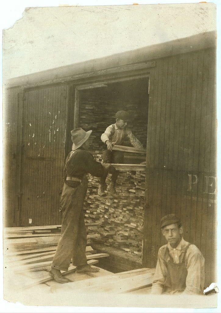 The image depicts a sepia-toned photograph that appears to be from the early 20th century, judging by its style and quality. It captures an outdoor scene with three individuals engaged in what seems like lumber-related work.

In the foreground on the right side of the photo is a young man seated against a wooden structure, possibly part of a barn or shed, wearing overalls indicative of manual labor attire from that era. His expression appears contemplative and he gazes directly at the camera with an intense look.

The central focus is two men standing in front of what looks like a large woodpile inside a covered area attached to a wooden structure. One man stands on higher ground within this space, reaching out as if handling or sorting through logs, wearing a hat for sun protection and casual work clothes. The second man is outside the enclosed area, stepping into it with one leg already in the opening, dressed similarly but without a hat.

The background features vertical wooden planks that form part of the wall enclosing this workspace, alongside stacks of lumber which suggest an industrial or rural setting related to woodwork or construction. No distinct branding or identifying marks are visible on any clothing or equipment, maintaining anonymity and focusing attention solely on their laborious task.