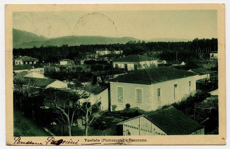 The image is a faded black-and-white photograph depicting an outdoor setting with rural architecture. In the foreground, there are several buildings that appear to be old and modest in size, possibly indicating historical or agrarian use. The central building has a simple rectangular shape with pitched roofing and visible windows. To its right stands another structure with similar architectural features.

In front of these structures is an area with trees, some small bushes, and what seems like agricultural plots with trellises supporting plants. There's also a pathway leading toward the buildings, suggesting regular access to this rural setting.

The background reveals more buildings scattered across hills or low mountains, indicating that the settlement extends beyond the immediate vicinity of these structures. The landscape is green and lush, hinting at fertile land possibly used for cultivation.

Atop one building on the right side stands a structure with an even smaller roof, which could be part of another utility shed or storage facility. A text inscription along the bottom edge provides context: "Pensione Panormita Tonfalo (PiETrASanta) ePanorama," hinting that this location may have been known as 'Tonfalo' and was likely a resting place, possibly for travelers.

The photograph has visible creases and aging marks suggesting it's an old postcard or print. The overal [...]