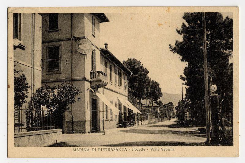 The image shows a black and white vintage postcard depicting an urban street scene from what appears to be the early 20th century. The setting is Marina di Pietrasanta, with buildings lining one side of a narrow road that curves gently out of view on the right-hand side. Trees are visible along the streetscape, providing some greenery and shade.

There's signage indicating "Marina di Pieterasanta - Focette - Viale Versilia." Architectural details include window shutters, awnings over windows or entrances, balconies with railings above ground level, and a sidewalk. A lamp post stands prominently on the corner of an intersection to the right side.

The image is worn around its edges, suggesting age and handling; some dirt marks are visible throughout. The overall atmosphere evokes a sense of quietness typical for a time period where such scenes were captured in everyday life before modern urbanization took over.