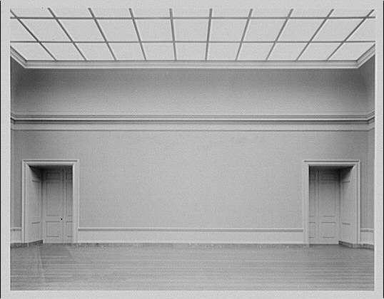 The image depicts an empty gallery room with a minimalist design, likely from the early to mid-20th century. The walls and ceiling are painted in a light shade of gray or white, creating a neutral backdrop for artworks. There are two doors on either side of the space, suggesting it's part of a larger building complex like an art museum.
The room features a wooden floor with visible lines of wood planks that run parallel to each other. The lighting appears natural and diffused, coming through large windows or possibly skylights in the ceiling grid pattern, which is common for galleries allowing sunlight to enhance artwork visibility without causing glare. There's no furniture or decorations within this space; it’s a blank canvas awaiting exhibits.
This photograph was taken by Horydczak Theodor (approx. 1890-1971), and its negative dimensions are approximately 8 x 10 inches, indicating that the original image might have been of professional quality or used for archival purposes in a gallery setting like National Gallery.
