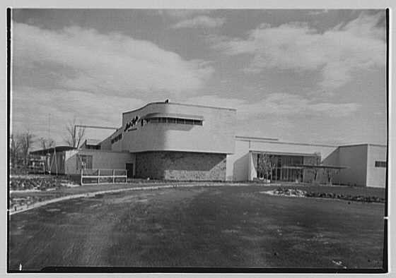 The image shows a black and white photograph of an architectural structure, which appears to be the exterior facade of a building from 1948. The architecture is modernist in style with clean lines, flat surfaces, and large windows that reflect minimalistic design principles popular during mid-20th-century America.
In the foreground, there's open space leading up to what seems like an entrance or access point for vehicles, as indicated by a paved area without visible markings. The building itself has multiple sections connected together with no clear roofline; instead, it features various horizontal and vertical planes that form its silhouette against the sky.

There are two main segments of the structure: one side is flat with large windows on top, possibly an atrium or lobby space given the height compared to other parts. The adjacent segment appears lower in elevation but maintains a similar design language with large openings for glass walls.
The photograph captures people near this part, suggesting it might be a gathering spot or entrance area within that section of the building.

There's no vegetation visible around the structure, indicating either early construction stages before landscaping took place or an urban setting. The sky is partly cloudy which contributes to the overall muted tones in the photo due to its black and white nature.
Overall, this image likely  [...]