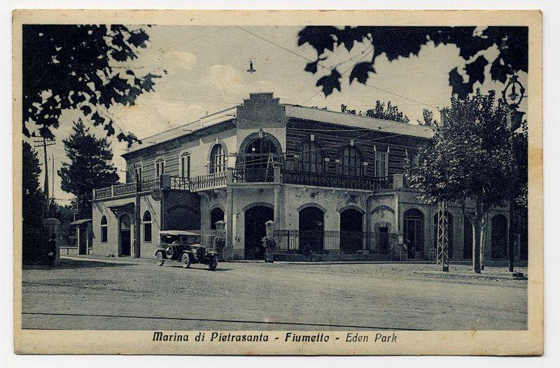 The image shows a historical black and white photograph of an old building labeled "Marina di Pietrasanta - Fiumetto - Eden Park." The architecture suggests European influence, with the structure featuring multiple levels, arched windows, balconies, and decorative ironwork on the second floor. A vintage automobile is parked in front of the building, indicating that this photo was taken during an earlier time period when automobiles were becoming more common but not yet ubiquitous.

The street appears quiet without much pedestrian activity or modern infrastructure. Trees are visible along one side of the road, suggesting some greenery around the area. The overall scene evokes a sense of nostalgia and gives insight into what urban landscapes might have looked like in an early 20th-century European town.