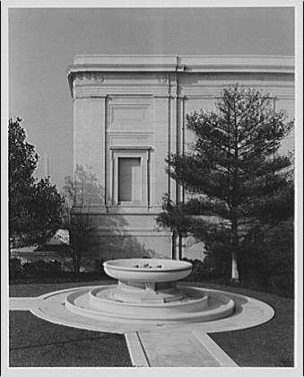 The image depicts a black and white photograph of the east corner of the National Gallery, showing an ornate fountain in front of it. The architecture of the building is classical with columns and detailed stonework on its facade. Two trees flank either side of the entrance to create symmetry around the structure. It appears to be from approximately 1920-1950 based on stylistic features, as indicated by a note stating "National Gallery ca. 1920-ca. 1950". Additional context or background information can potentially be found in National Gallery of Art archives for more insights into the period and significance of this image.