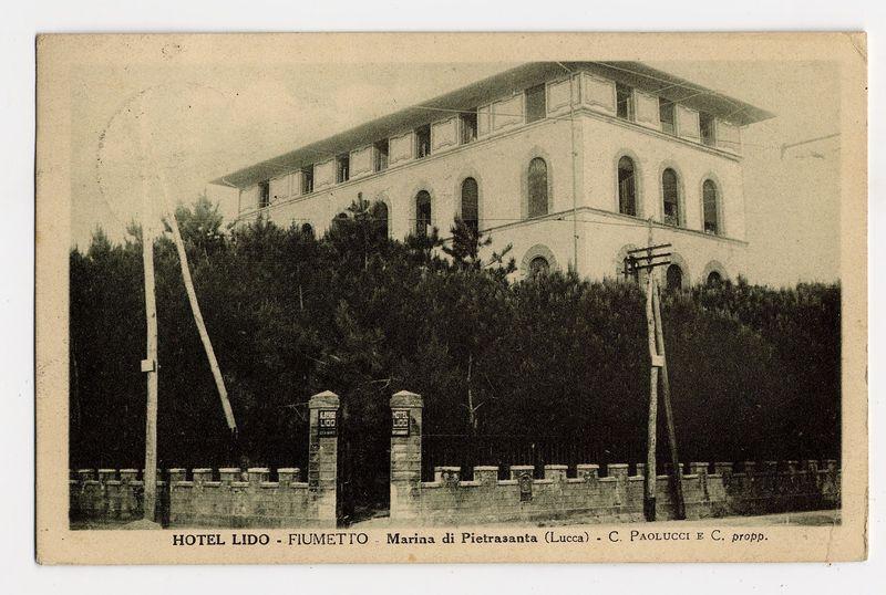 The image displays an old black-and-white photograph of a two-story building, labeled as Hotel Lido - Fiumento. It is situated at Marina di Pietrasanta in Lucca and credited to C. Paolucci E.C., likely indicating the photographer or studio that produced it. The structure appears stately with multiple arched windows on both levels and what seems like balconies overlooking a tree-lined area, possibly suggesting an early 20th-century architectural style.

In front of the hotel is a gated entrance marked "HOTEL LIDO" in large letters at two distinct locations—a pair of stone pillars flanking the gate. The photograph has signs of wear with creases and some discoloration over time, indicating it may be an antique or very old postcard that could have been used for travel promotion during its prime.

The image is bordered by a thin white margin which suggests it was once part of a larger collection like a scrapbook or album. There are no people visible in the photograph, keeping the focus solely on the architectural subject and the surrounding environment.