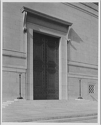 The image depicts a large, ornate main door of an institutional building, likely from the early 20th century. The photograph is in black and white, suggesting it might be historical or archival material. The doorway features grand stone columns on either side with decorative capitals, supporting a substantial lintel above the entrance.

On each corner post beside the doors are slender lampposts mounted securely to the stonework of the building facade. These posts appear functional as well as aesthetic elements in this architectural design. Below and around the door, two small ornamental water spouts direct rainwater away from the foundation on both sides.
The National Gallery is likely an art museum or similar cultural institution based on its imposing entrance and classical architecture style reminiscent of neoclassicism prevalent during that era.