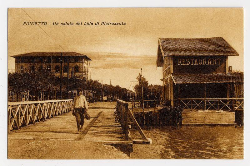 The image depicts a sepia-toned vintage photograph, suggesting it is from an earlier time period. It shows a person walking on what appears to be a boardwalk or pier over water towards the left side of the photo where another structure can be seen in the background. The individual seems casually dressed and carries something in their hand which could possibly be bags or groceries.

On the right, there is a prominent building labeled "RESTAURANT," indicating that it was once likely a dining establishment open to patrons visiting this waterfront area. Overhead text reads "FIUMETTO - Un saluto del Lido di Pietrasanta" translating from Italian as "Fiumetto - A salute of the Lido of Pietrasanta." This suggests the location is near or in Pietrasanta, which could be a town known for its seaside attractions.

The water surface reflects some structures and light, hinting at an open setting with ample sunlight. The overall atmosphere appears calm and leisurely, typical of a day at a waterfront recreational area during that era.