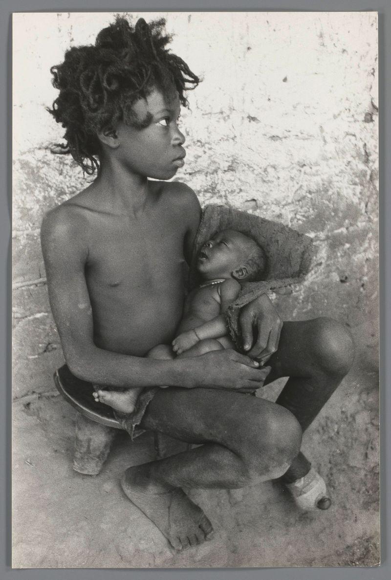 The image depicts a young child with curly hair, sitting on the ground. The child is shirtless and appears to be in a modest environment suggested by the rough texture of the wall behind them. On their lap rests another small figure, likely an infant or toddler also without clothing, suggesting they might not have access to basic needs such as proper attire.

The photograph has a monochromatic tone with varying shades from white to black and is framed within what looks like a dark border typical for printed photographs of the era. The visual style hints at it being taken during mid-20th century Europe, possibly in post-war context given its stark simplicity and the lack of modern amenities.

There's an element of vulnerability conveyed by both figures; their bare feet, simple surroundings hinting towards poverty or hardship they might be facing. No explicit action is captured but a sense of quiet endurance is palpable through their stillness against what appears to be a nondescript background devoid of any distinctive objects that could offer additional context.

The absence of color and the grainy texture suggest it may have been taken with an analog camera, likely during or shortly after World War II when many European nations were under post-war reconstruction.