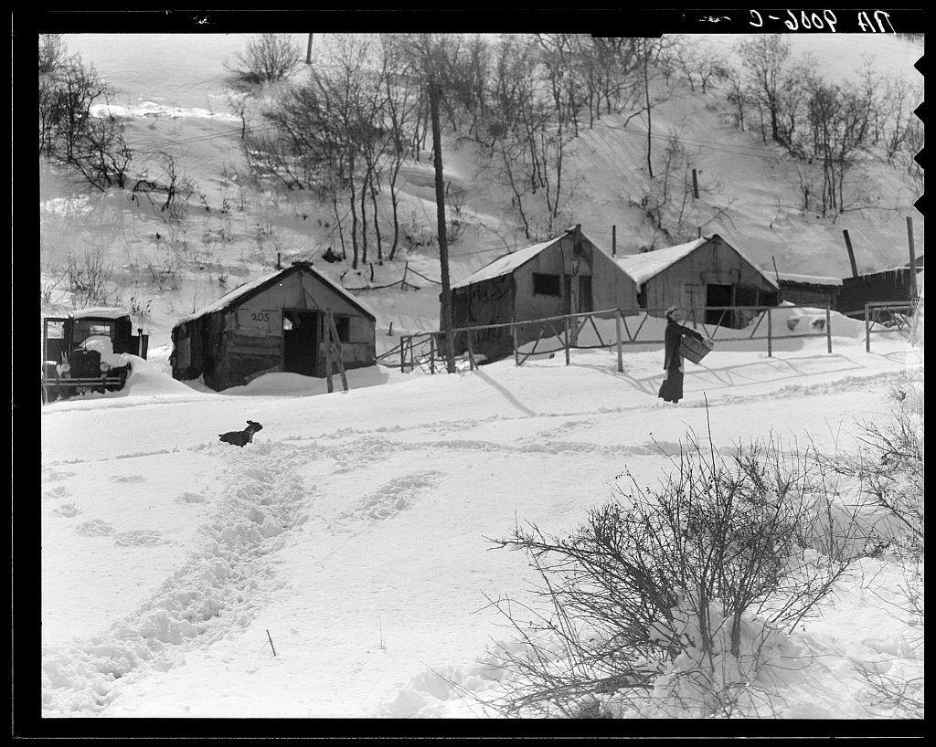 The image depicts a snowy rural landscape with rustic buildings, including several small wooden structures and sheds. A single person is visible in the scene holding what appears to be a bundle of firewood or logs. The terrain features undulating hills blanketed with snow, suggesting cold weather conditions typical for winter months.

In the foreground, there's an animal lying on the ground near tracks carved into the snowy surface by foot traffic. Bare bushes and leafless trees are scattered throughout the scene, emphasizing the wintery atmosphere. A vehicle is partially visible at the left edge of the photo, reinforcing the notion that this environment might be associated with manual labor or transportation in challenging conditions.

The black-and-white nature of the photograph adds a historical feel to the setting, hinting it could have been taken several decades ago when such rural and industrialized landscapes were more common.