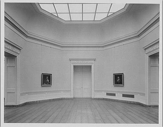 This black and white photograph displays an empty gallery room, likely from the early 20th century. The high ceilings are accentuated by a large skylight that allows ample natural light to flood into the space. On each wall of this well-lit environment hangs one portrait within its own ornate frame; these artworks seem to be centrally positioned along the walls for optimal visibility.

The room itself is spacious, with hardwood flooring and decorative baseboards running horizontally around all four sides. The far end features a closed door leading presumably into another section or possibly an exit from this gallery space. In front of one wall lies what appears to be a small heating vent, indicative of older building infrastructure that necessitated individual climate control in such large spaces.

The overall atmosphere is quiet and studious; it seems designed for the purpose of art exhibition rather than social gatherings. It evokes an ambiance conducive to contemplation or study of artworks on display without distractions from other patrons or loud noises within this serene space.