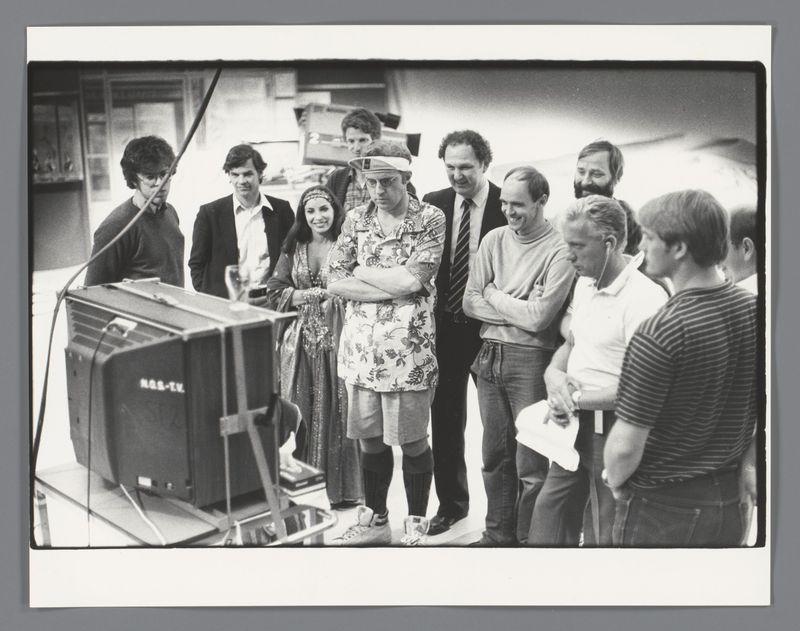 A group of eleven people gathered around a vintage portable television with "HOS-TV" labeled on it. The setting appears to be an indoor space, possibly part of the backstage area or production office for a media event such as a comedy show given the context provided by the reference image link.

The individuals display varied expressions and postures; some are attentive while others exhibit smiles or engaged in conversation with one another. Among them is someone wearing what looks like a novelty hat over their head, possibly indicating a role of importance within this group dynamic.

Behind the central figures is an assortment of equipment indicative of media production such as cables and other devices. The overall atmosphere suggests anticipation for something on display through the television or that they're about to engage in some form of performance related activity.