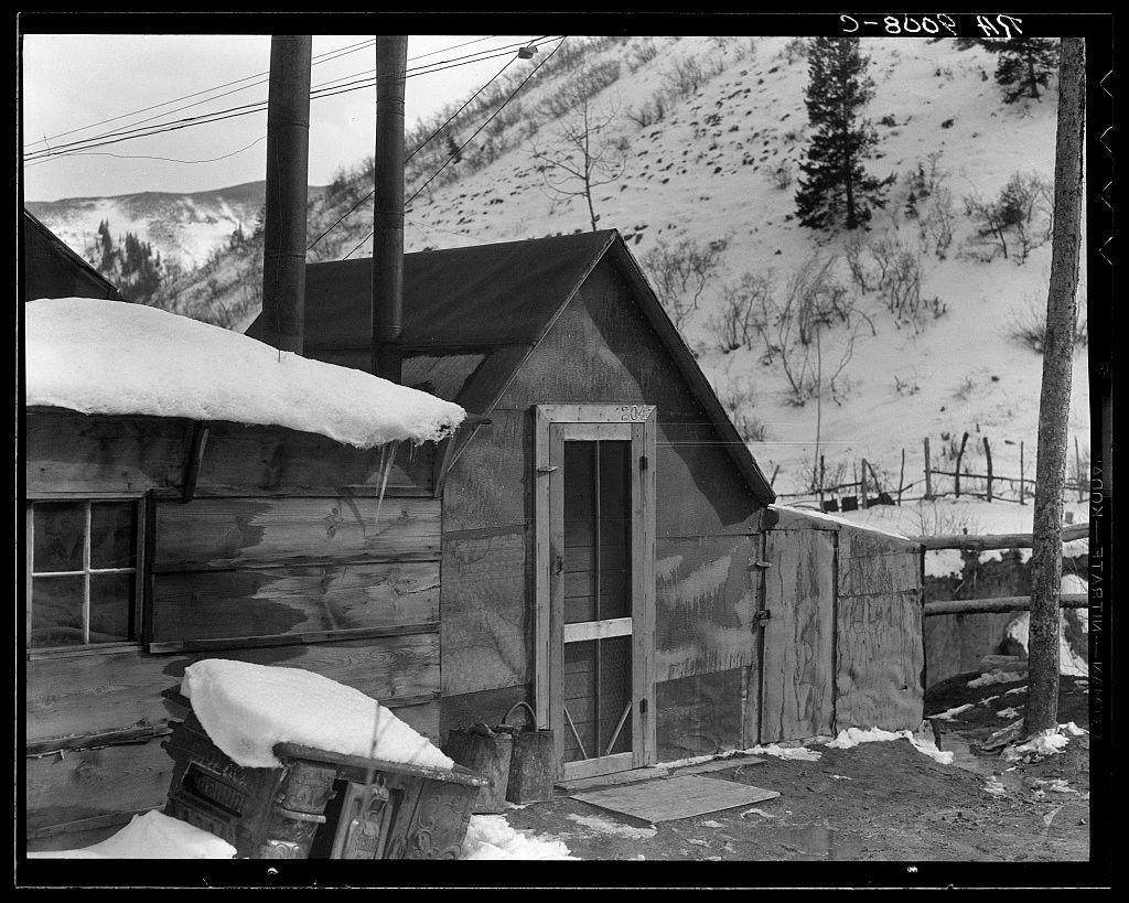 A black and white photograph of a small wooden cabin surrounded by snow. The structure appears aged with weathered wood, particularly noticeable on the front door which has large glass panes in need of repair or replacement as they are cracked and missing pieces. Snow accumulates on top of a pile of items outside including what seems to be an old stove covered partially with snow, suggesting cold temperatures likely during winter months.

The cabin is marked "804" above the front door entrance which may indicate either its house number or perhaps it's part of a larger compound that isn't fully captured in this photograph. There are visible power lines running along one side and another structure can be seen partially behind, with trees scattered across the snowy hillside.

The absence of people provides an air of solitude but suggests resilience against harsh conditions as evidenced by the cabin’s ongoing use despite its age or wear-and-tear. The surrounding landscape is barren except for evergreen trees that have endured the cold without losing their foliage suggesting a winter season where snowfall has occurred, typical in regions near mining towns like Price, Utah which was known to be associated with coal mining activities as indicated by the photograph’s caption.

This image captures not just an isolated building but tells a story of endurance and possibly hardship o [...]