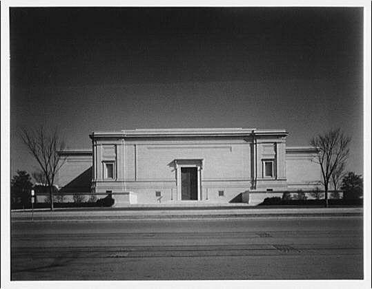 The image is a black and white photograph of the west end facade of a classical-style building, likely taken during the early to mid-20th century. The structure has symmetrical architecture with rectangular shapes and features such as pilasters, a cornice at the top edge, and large double doors centered on its front façade.
There are no visible signs or identifying marks that explicitly indicate what this building is meant for; however, it resembles institutional buildings like museums or libraries from that era. The sky appears clear with ample sunlight casting shadows on the ground in front of the building. Two leafless trees flank either side of the structure, suggesting a barren season such as winter.
The photograph has no discernible people or moving objects within its frame, which focuses solely on capturing the building's architectural details and setting it against an open road that runs parallel to it without any visible traffic.