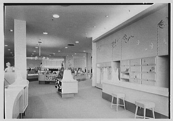 The image depicts an interior view of a department store, likely from the early 1950s based on its design and presentation. The setting appears to be a cosmetics or perfume section with various displays showcasing products such as bottles and boxes arranged neatly behind counters. Above these display cases are overhead signs that seem to indicate different sections for items like eyeshadows or lipsticks, though specific labels aren't clearly visible.

The store has a modernistic design with clean lines, featuring white flooring and walls which create an airy atmosphere. There's also a seating area at the front of the image where customers might wait while shopping or resting. The lighting is bright and evenly distributed across the space via ceiling-mounted fixtures. Overall, this establishment seems to be well-maintained and organized for customer convenience during its time period.