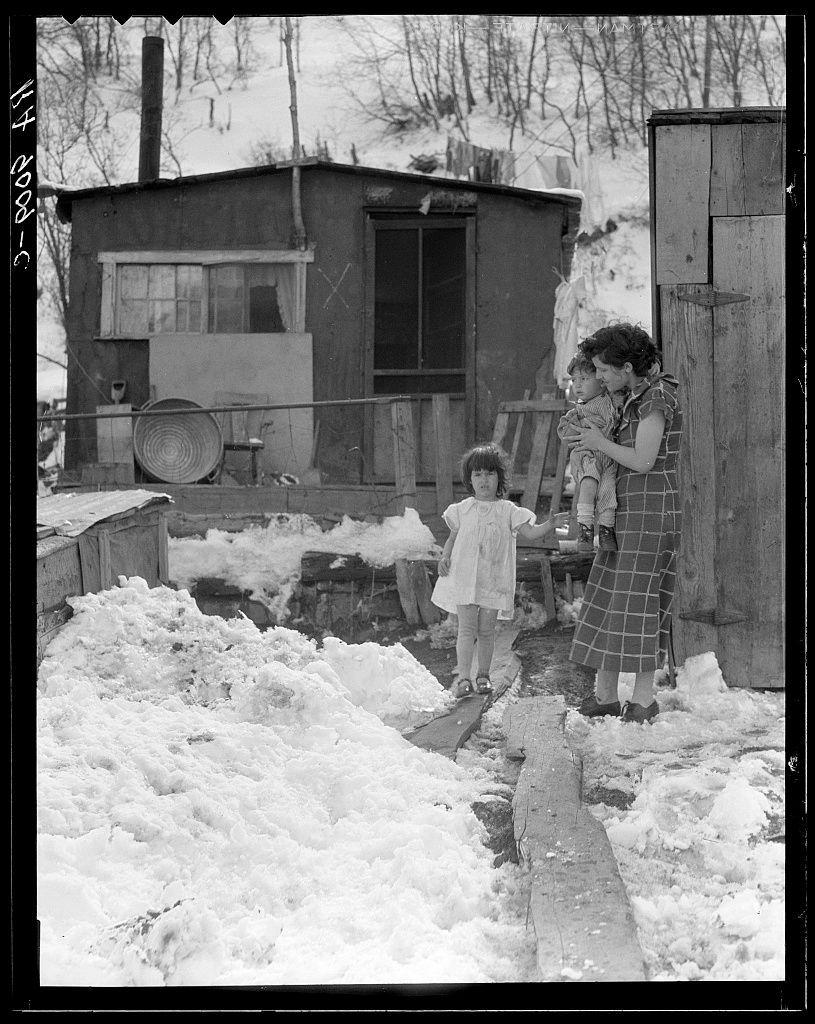 The image depicts an outdoor scene with three individuals during winter. In the foreground, there's a young child in a white dress standing on what seems to be snow-covered ground and reaching out for support from a wooden plank that is placed across two logs or beams amidst the piled-up snow. This individual appears to have unkempt hair and wear minimal clothing suitable for cold weather.

Next to this child stands an adult woman, likely their mother, holding another young child wrapped in what looks like layers of cloth, possibly providing warmth against the chilly conditions. The older woman is dressed in a checkered dress with dark sleeves and shoes that are partly covered by snow, suggesting they've been outdoors for some time.

Behind them rises a modest wooden shack or small house structure characterized by its simple construction, single window without curtains, an open door to what could be a porch area, and various items scattered around including a large metal bowl on the railing of the porch. The environment is rural with bare trees in the background indicating it's likely late winter or early spring.

The ground outside shows significant snow accumulation suggesting heavy recent snowfall, which adds a stark contrast against the dark color of the shack. There are no visible signs that indicate modern amenities; instead, this setting evokes an impression of rus [...]