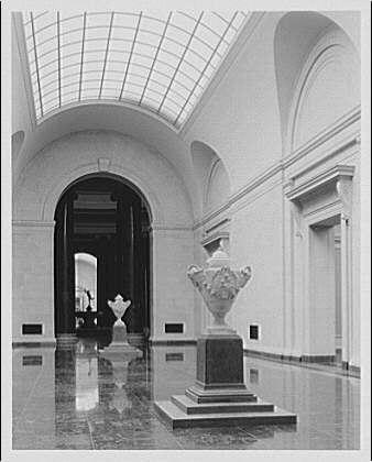 The image depicts a black and white photograph of an elegant, spacious hall with high ceilings made up of arched windows allowing sunlight to stream in. The architecture is grandiose, featuring tall columns, ornate decorations on the walls, and large marble floors reflecting light from above.

In the center of the frame are two prominent urns placed symmetrically against a wall; these classical-looking vases appear to be made of white stone or marble with intricate carvings. The room gives off an atmosphere reminiscent of museum interiors due to its refined decor and reflective surfaces, suggesting that this could be part of a gallery displaying art pieces.

Additional information about the image indicates it is associated with Horydczak (Theodor) whose work dates approximately from 1890-1971. The photograph was taken on one negative measuring safety size at eight inches by ten inches and can be found in National Gallery of Art, specifically within East Hall featuring monumental urns attributed to Clodion circa 1920-ca. 1950.

The photo captures a striking contrast between the natural light entering through the windows and the artificial ambient lighting illuminating this stately space filled with historical artifacts or decorative pieces. The reflection on the polished marble floors adds depth, emphasizing both the scale of these urns as well as the architectural beauty  [...]