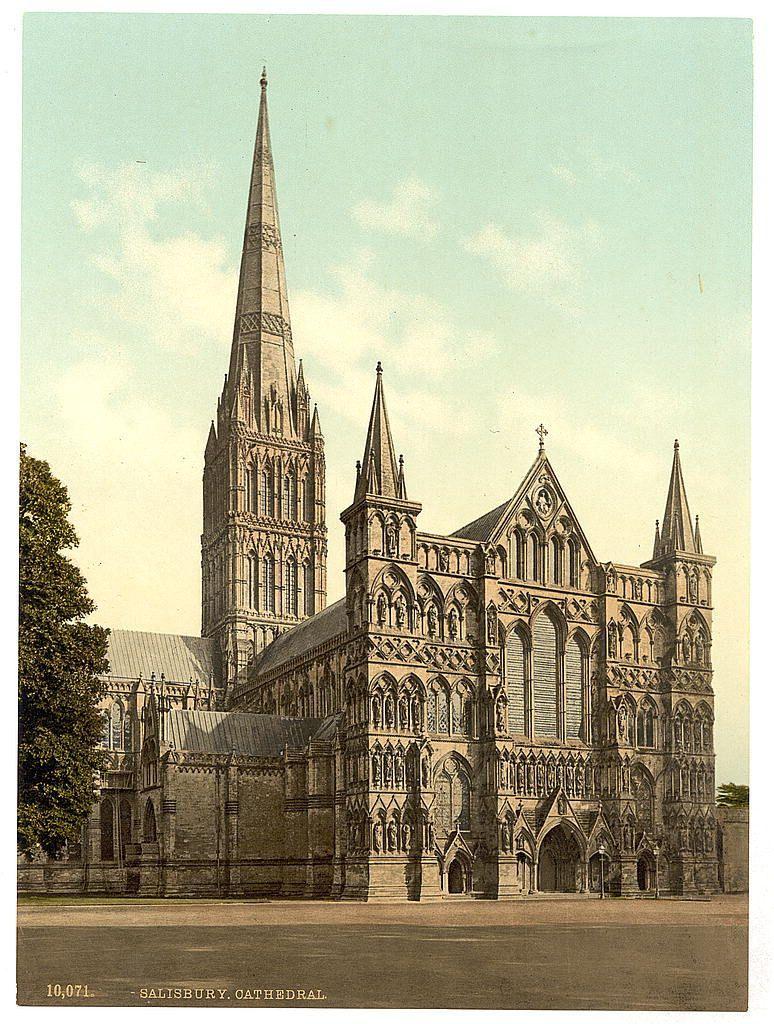 The image depicts a grand Gothic cathedral with intricate stone carvings and towering spires. The central feature is the slender, pointed spire that rises prominently against a partly cloudy sky. To its right stands another tower topped with an ornate cross, indicative of religious significance. Multiple buttresses support the large facade adorned with rose windows, lancet windows, and flying buttresses typical of Gothic architecture. A well-maintained square is in front of the cathedral, suggesting it may be a public or significant space within its locale. The overall scene conveys historical grandeur and architectural elegance.