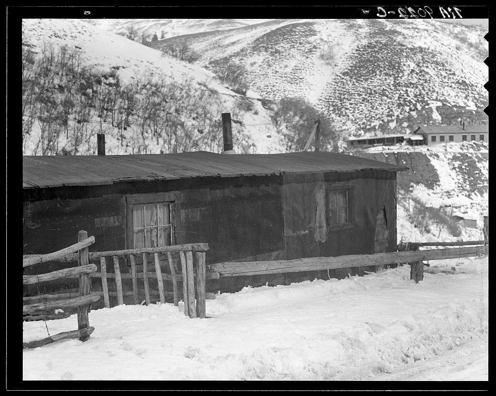 The image depicts a black and white photograph of an old wooden cabin nestled in a snowy landscape. The cabin, showing signs of wear with its darkened exterior walls and boarded-up windows, is surrounded by snow-covered ground that extends into the distance, indicating heavy winter conditions.

In front of the cabin stands a rustic wood fence partially buried under the snow, suggesting recent or ongoing snowfall. The terrain beyond features rolling hills blanketed in patches of snow against barren trees with no foliage visible due to the cold season.

The backdrop reveals more structures further up on the hillside, which could be additional housing units associated with the cabin shown. A power line is also seen running diagonally across the frame from top right to bottom left corner behind the main structure, hinting at a connection to electricity or telecommunications services in this remote setting.

This image captures an atmospheric scene of isolation and hardship typically found within mining towns like those depicted by Dorothea Lange during her work documenting rural life.