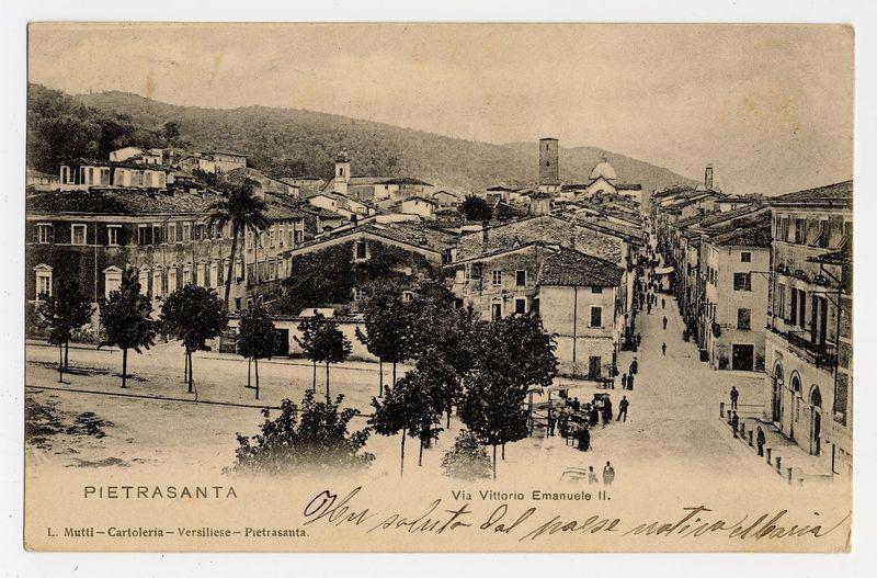 The image is a historical black-and-white photograph depicting an old European town, specifically Pietrasanta via Vittorio Emanuele II. The street appears wide with several buildings on both sides, featuring pitched roofs and various architectural styles indicative of the late 19th or early 20th century.

On one side of the street stands a prominent church with visible bell towers rising above other structures. Trees line parts of the sidewalk alongside cobblestone streets where horse-drawn carriages are parked beside buildings adorned with classical facades and shuttered windows, suggesting residential use.

Pedestrians are present on the sidewalks, some walking in groups while others stroll alone or engage in conversation. The environment is bathed in a muted light that suggests either dawn or dusk due to its softness and angle of illumination casting long shadows across the scene.

The overall atmosphere evokes a sense of tranquility but also hints at an active community life from another era, characterized by slower-paced movements and interactions among people gathered on the streets.