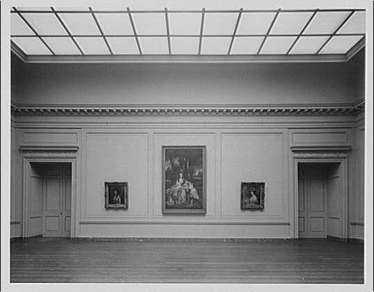 The image depicts an exhibit room within the National Gallery of Art, as indicated by a reference to its exhibition period between 1920 and 1950. The photograph is black-and-white, suggesting it was taken during that time frame or later in the mid-20th century when color photography became more prevalent.

The museum space features white walls with ornate molding at regular intervals around the perimeter of the room. Two doorways flank opposite sides, each leading into a hallway beyond which one can imagine other gallery spaces extending outwards from this central area.

Three large framed paintings are mounted on two parallel wall sections within view: On the left side is an image too indistinct to describe but appears darker and possibly monochromatic; in between these doors hangs what seems like a group portrait featuring multiple figures, likely historical or classical subjects common in such institutions; finally, on the right lies another large framed painting depicting perhaps a solitary figure.

The ceiling has frosted glass blocks which allow diffused daylight into this indoor space. The flooring is polished wood planks that reflect light subtly and enhance the room's elegant atmosphere. This particular exhibit was captured by photographer Theodore Horydczak (1890-1971), according to metadata provided, who likely documented various aspects of art exhibitions duri [...]