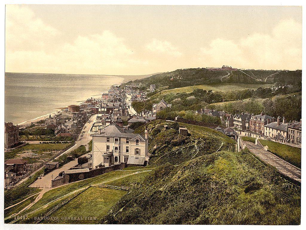 The image displays a historical view from an elevated perspective overlooking a coastal town. The scene captures the curvature of a seafront, dotted with rows of terraced houses and buildings extending into the distance along what appears to be a promenade or beachside walkway. A prominent hill rises on one side, adorned with greenery and some structures atop its crest, including possibly a lighthouse or tower near the summit.

In front of this town backdrop lies an expanse of grassy slopes leading down towards the seafront below. The foreground shows winding pathways cutting through lush vegetation, suggesting parks or gardens within the urban setting. A railway track runs alongside these paths on what seems to be embankments or elevated tracks, hinting at a transportation route for trains.

The overall coloration and texture of the image suggest it is an old photograph or postcard, with sepia tones dominating much of the scene except for certain highlights where natural light reflects off buildings. The weather appears fair as indicated by the visibility across the landscape and partly cloudy skies above. A caption at the bottom indicates this was a general view from Sandgate in England during approximately 1890 to 1900, providing historical context to the image's location and period of capture.