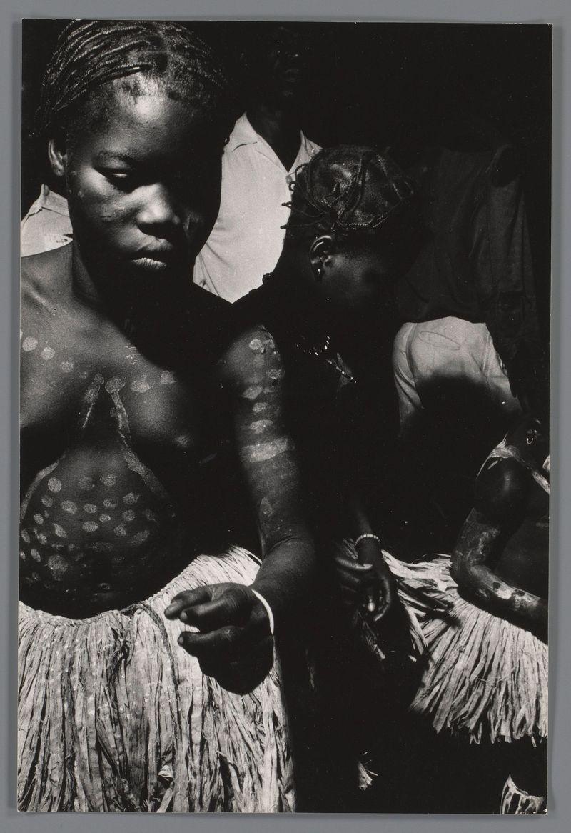 The image is a black and white photograph featuring two individuals, likely taken in the context of cultural or traditional practices. The person on the left appears to be adorned with body paint that includes circular patterns over their chest area and arm tattoos, suggesting some form of ceremonial significance. They are wearing what seems like parts of traditional clothing made from rough-hewn fabric strips, commonly known as a 'hula skirt'. Their expression is somber or introspective.

The individual on the right appears to be in mid-motion with an outstretched arm, possibly reaching for something outside the frame. This person also wears a similar style of dress and has their arms adorned with what could be additional body paint or jewelry. The background includes partial figures wearing light-colored clothing, indicating more people are present but not focused on.

This photograph is captioned "Kinderen genaamd 'Ganza's' tijdens dansplechtigheid na afloop van een besnijdenis," which translates to "Children named 'Ganza's' during the quietude after a haircut." The image, taken by Ed van der Elsken in 1956-1957, likely captures moments from an event or ritual involving community members.