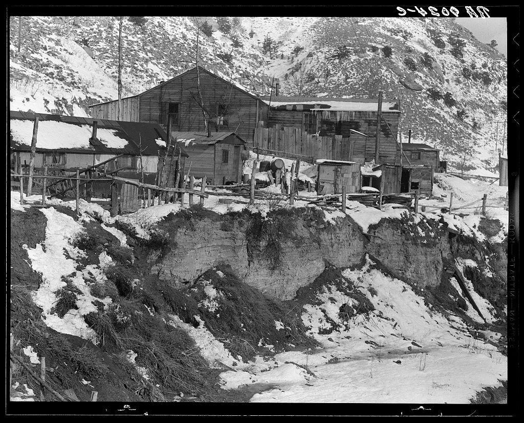 The image is a black and white photograph depicting an old mining town with dilapidated buildings on the side of a snowy hillside. The structures appear to be made from wood, showing signs of weathering and abandonment. Snow covers parts of the landscape around the homes, suggesting cold or winter conditions. There are fences, barrels, and what looks like remnants of wooden planks scattered throughout the scene. No people are visible in the photograph, adding a sense of desolation. The text "5-1908 3D" is printed on top right corner, possibly indicating a date stamp from an archive or collection where this photo resides.