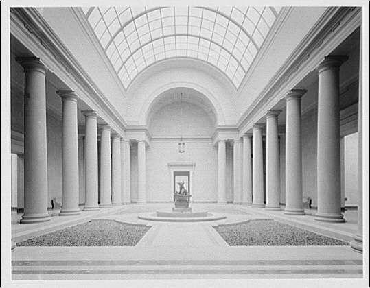 The image is a black and white photograph depicting the interior of a grand, neoclassical structure with an expansive central courtyard. The architecture features tall columns supporting a curved archway that leads to a skylight ceiling, allowing natural light to illuminate the space. In the center stands a statue on a pedestal, surrounded by two circular flower beds filled with what appears to be gravel or small stones.

The room is symmetrical and has an elegant, austere beauty typical of neoclassical design elements from around 1920-50. The absence of color in the image emphasizes the geometric shapes and contrasts between light and shadow within the space.

This architectural photograph captures a historical moment at what seems to be part of a museum or gallery, possibly showcasing its importance as an art institution during that period.