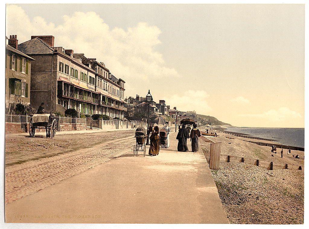 The image depicts a vintage scene, likely from late 19th to early 20th century England. A group of people is strolling along what appears to be a promenade by the seaside. They are dressed in long dresses and bonnets typical for that era's women's fashion. The attire suggests they may be on vacation or enjoying leisure time, as such clothing was not commonly worn outside home environments during this period.

The setting includes multi-story buildings with several floors of windows, indicative of a town rather than rural surroundings. These could potentially serve various purposes but are most likely residential in nature given their proximity to the promenade and beach.

A horse-drawn cart is visible on the left side of the path, adding to the historical ambiance. The sandy area adjacent to the promenade suggests that this part of Sandgate was a popular destination for seaside activities during its time.

The sky appears partly cloudy with enough sunshine suggesting it's not too cold or overcast. The beach extends into the distance where fewer people are visible near the waterline, which might indicate low tide or simply less crowded areas further from the main promenade path.

Overall, this image captures a moment of calm and leisure in an English seaside town during its heyday as a vacation destination.