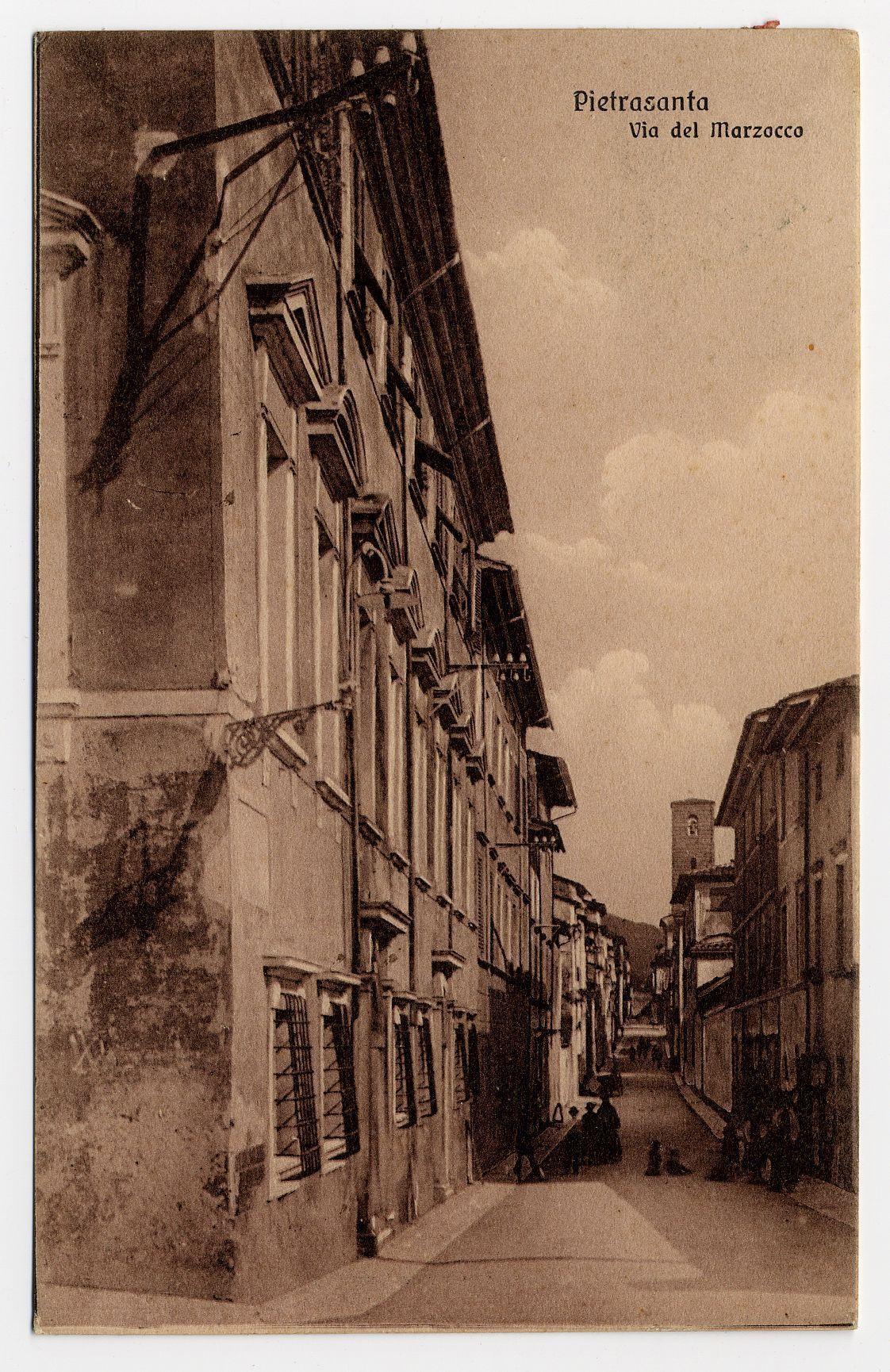 The image depicts a sepia-toned photograph of an urban street scene, likely taken in the late 19th or early 20th century. The architecture is characterized by tall buildings with ornate facades and large windows, indicative of European style possibly found in Italy given the reference to Pietrasanta. Some balconies have bars attached at lower levels while higher sections are accessible without barriers.

The street appears narrow but seems busy despite a lack of visible people or vehicles due to the vintage quality of the image which could mean it's either a still from motion pictures, such as early cinema with very few moving subjects captured in each frame. The sky is cloudy and gray, suggesting an overcast day.

Atop one building on the right side stands what appears to be part of a clock tower or steeple, further emphasizing this image could represent European architecture rather than American styles which typically lack such ornamental towers without practical purpose like housing bells or utilities in non-religious contexts. The presence of utility lines attached to buildings is visible.

The photograph shows signs of age with slight creases and some discoloration on the paper, giving it a historical feel. There's text at the top reading "Pietrasanta Via del Marzocco," which identifies the location as Pietrasanta street via del Marzocco in what could be Pietrasanta  [...]