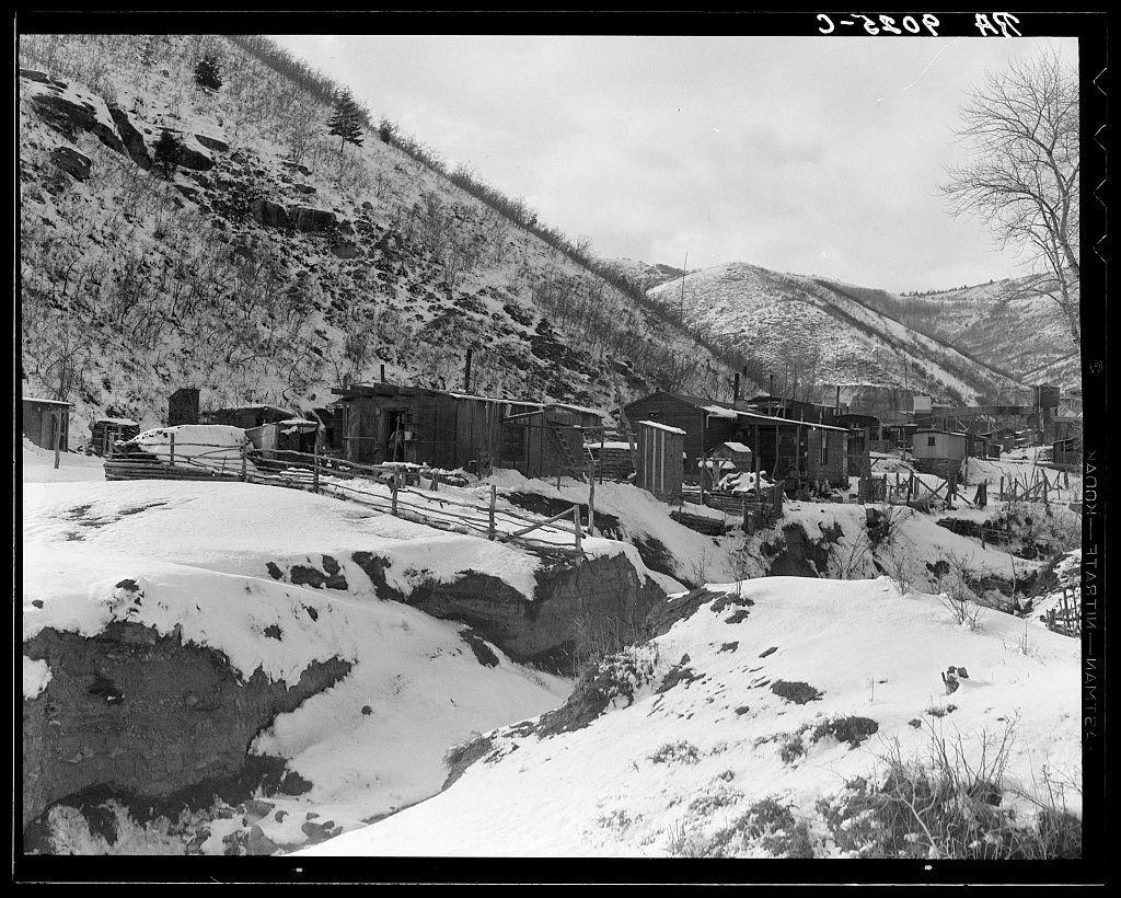 The image is a black and white photograph depicting an old mining town situated in a snowy, mountainous landscape. The scene shows several small wooden structures with corrugated metal roofs scattered across the hillside. These buildings appear to be hastily constructed homes or cabins typical of early miners' dwellings from the late 19th century.

The terrain is rugged and covered partially by snow, indicating cold weather conditions likely during winter months. Tracks in the snow suggest recent activity around these structures. The surrounding hills are barren with sparse vegetation, highlighting a harsh environment suitable for mining but challenging to sustain habitation.

A wooden fence runs along part of the property line between some of the buildings, adding another layer of rustic charm to this historical setting. In the foreground on the left side, there's an exposed earth mound covered partially by snow, possibly indicating erosion or land disturbance due to mining activities.

The sky is overcast with a mix of clouds and clear patches, suggesting variable weather conditions typical for mountainous regions during winter seasons. The overall atmosphere conveyed by this image reflects resilience amidst tough living conditions in the early days of coal mining communities like those found in Utah's Price area.