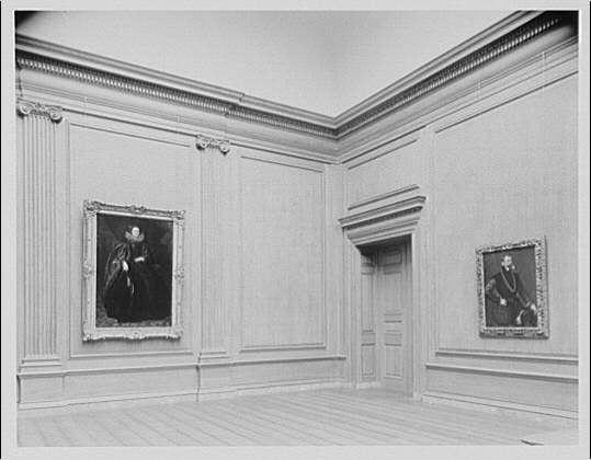 The image depicts a classic-style interior room, likely within an art gallery or museum based on its design and context. Two framed portraits hang on the walls; one to the left side featuring what appears to be a historical figure in darker attire with white ruffled clothing around their neck, possibly from an earlier era such as the 17th century due to the fashion depicted.

The second portrait is smaller and situated near the right wall, portraying another individual. The room has wood-paneled walls and floors, suggesting a formal or prestigious setting often associated with classical art displays. Overhead molding adds to the stately ambiance of this space. A single door can be seen in the center of one side of the room.

There is no visible activity besides these static elements within the image; it captures an atmosphere rather than dynamic movement or interaction between subjects, which aligns well with a historical context likely during late 19th to early 20th centuries based on architectural style and monochrome photography.