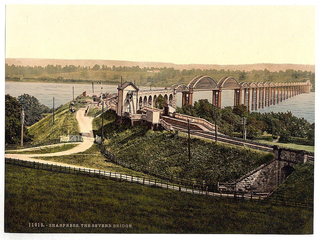 The image depicts a vintage photograph or postcard showing an expansive view of the Severn Bridge in Sharpness, England. The bridge spans across a river with its distinctive structure highlighted by numerous arches and supports that allow for railway tracks to pass over it. To one side is an elevated roadway leading up to the bridge's entrance, featuring two imposing towers connected by a series of staircases or ramps, suggesting accessibility features typical of bridges from this era. The surrounding landscape includes rolling green hills with patches of grassy fields, and there appears to be fencing alongside pathways on both sides of the image. A distant riverbank is visible across the bridge's length, hinting at its location as a significant waterway crossing.

The photograph has an old-fashioned aesthetic, characterized by faded colors that range from sepia tones near the buildings to muted greens in the foliage and grays in the man-made structures like staircases or roads. Notably, there is text on the bottom left of the image stating "1015 - SHARPNESS THE SEVERN BRIDGE," which likely serves as a caption indicating both its location and subject matter.

This historical representation provides insight into early 20th-century infrastructure engineering, showcasing how bridges were designed to facilitate transportation across rivers before modern vehicular spans. The p [...]