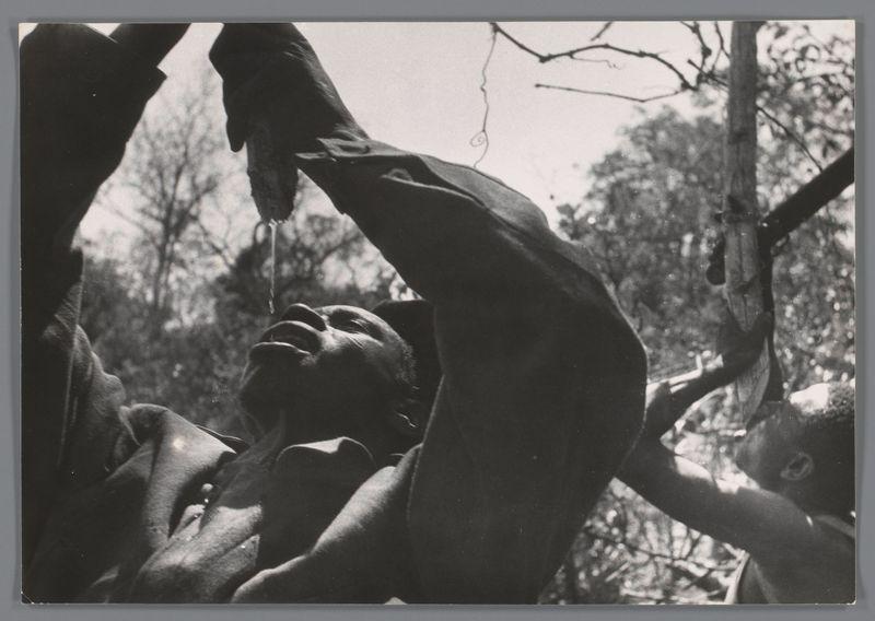 The image depicts a group of people gathered closely together, participating in an outdoor activity. They appear to be sitting on the ground with their knees drawn up and legs crossed at the ankles. Their bodies are oriented towards each other, suggesting that they might be engaged in conversation or some form of interaction.

One individual stands out prominently as he is captured pouring a liquid from one cup into another. This action implies social drinking, possibly involving beer given its traditional association with such gatherings. The person's attire consists of dark clothing which contrasts against the lighter background of trees and foliage.