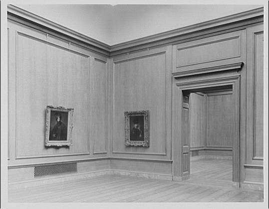 The image is a black and white photograph of an empty room, which appears to be part of an art gallery or museum. The walls are paneled with wood wainscoting extending halfway up the wall height. Two framed portraits hang on the left side; one portrait depicts what seems like a seated figure in formal attire, while the other portrays another individual possibly from behind.

On the right-hand side stands open doorway leading to an adjoining room or hallway within the gallery space. The flooring consists of wooden planks that match the walls' color and texture. A small heating register is visible on one wall near the floor level at a lower height than typical, indicating older infrastructure systems in use during this period.

The image has a classic feel with its composition reminiscent of early 20th-century interior design. The absence of people or modern elements suggests that it might be an exhibit room within National Gallery III from approximately between ca.1920 to ca.1950, as indicated by the text below the photograph.

The image is credited to Horydczak, a photographer active around 1890-1971 and associated with Theodor in some capacity (possibly indicating that it was taken or owned by someone named Theodore). This credit appears alongside an identification code "691e" which could be used for cataloging the photograph within archives.

The source of this image is [...]