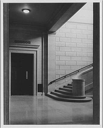 the image displays a black and white photograph of an interior space, specifically the ground floor stairway within what appears to be a museum or gallery. The architecture is classic with tall ceilings and large windows allowing ample light into the corridor.
In the foreground, there's a set of stairs leading up from the entrance level where two doors are visible; one seems like it leads outside while another could possibly connect to other levels. To the right side, we see an elevator marked "Elevator" above its door which is closed and has round buttons for floor selection.
The main focus of this image though lies in a curved stairway leading upwards on the left-hand side from where one can infer that there are additional floors beyond view within this establishment. The flooring appears to be polished concrete or stone, reflecting some light off their surface creating highlights across them.
A trashcan stands alone at an entrance near these stairs suggesting it serves both as functional and aesthetic purposes in maintaining cleanliness while adding a touch of modernity to the otherwise historical setting.

The lighting is subtle yet effective highlighting key architectural features such as windows, doors, elevator access point etc. The overall atmosphere exudes calmness with no visible human presence which adds an element of solitude or quietude possibly hinting at la [...]