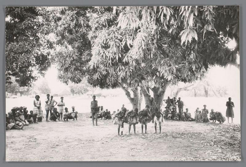 The image is a black and white photograph depicting an outdoor gathering of people in what appears to be rural or tribal setting. A large tree with dense foliage provides shade for the group, which consists predominantly of individuals dressed in traditional clothing. Some are standing while others sit on the ground, creating a semi-circle around two central figures who appear engaged in conversation or performance; one is holding what looks like a basket.

The attire varies among the participants: some wear simple dresses and wraps indicative of local fashion, suggesting cultural diversity within this community. The people exhibit relaxed postures, with hands resting at their sides, on knees, or folded over laps, indicating comfort and familiarity in each other's company. There is also an array of activities occurring simultaneously; individuals are standing by the tree, some looking towards a person who stands prominently holding something up for display.

The environment includes leaf litter scattered across the ground, hinting at open-air conditions typical of rural settings or outdoor communal spaces. The background is softly focused but shows more trees and possibly bushes, reinforcing the natural ambiance. There's no clear indication of modern infrastructure like roads or buildings, suggesting a setting removed from urban development.

Overall, this scene captures  [...]