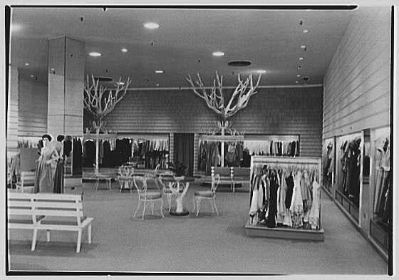 The image is a black and white photograph depicting an indoor retail environment, specifically within a clothing store. It shows two women standing in the middle of the space; one appears to be browsing clothes while another stands nearby conversing with her.

The interior features several elements indicative of mid-20th-century design: large metal-framed mirrors line the walls and hang from ceilings, reflecting parts of the shop's contents. There are also decorative tree branches without leaves arranged as a form of display in the center area, likely serving both an aesthetic purpose and to demarcate different sections within the store.

Benches for seating can be seen on one side, suggesting that customers might wait or rest while shopping. Clothing racks filled with garments stand prominently throughout the space; some are placed near walls displaying hanging outfits, while others are arranged in central areas for easy access by shoppers.

The lighting appears to come from overhead fixtures and possibly natural light coming through windows not visible within this frame of the photo. The overall atmosphere is that of a clean, well-maintained shopping area with an organized layout meant for customers' comfort and ease as they browse merchandise.