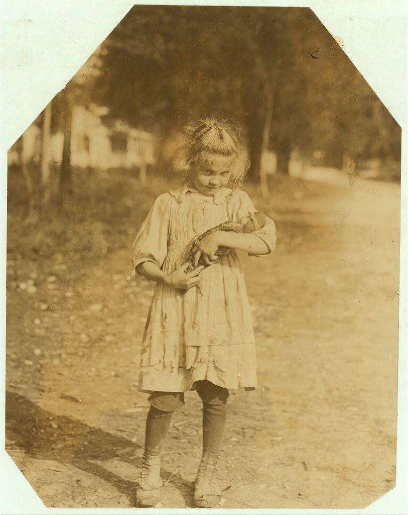 The image depicts a young girl in an outdoor setting during what appears to be late 19th or early 20th century. She is dressed in period-appropriate attire, consisting of a long-sleeved dress with ruffled cuffs and hem, knee-length stockings, and lace-up boots that reach mid-calf length. The fabric has a textured appearance indicative of the era's clothing materials.

The girl stands on an unpaved path or road surrounded by vegetation, suggesting a rural or semi-rural environment. She holds what seems to be a small bird in her hands with both arms. Her expression is not clearly discernible due to privacy reasons but appears attentive and focused on the creature she is holding.

This sepia-toned photograph has an octagonal border typical of albumen silver prints from that time period, which were commonly used for portrait photography before glass plate negatives became standard in the mid-1850s. The image carries a sense of innocence and simplicity reflective of childhood during those times.