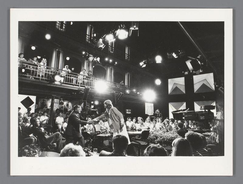 The image depicts a lively scene from what appears to be a television talk show set in an indoor venue, possibly during the 1970s or early '80s judging by the style of clothing and black-and-white photography. A central figure stands on stage addressing a seated audience while being filmed with professional lighting equipment overhead. The individual is dressed casually yet flamboyantly, wearing a striped top and dark pants. Surrounding this person are various other individuals: some standing in what seems like an entrance or balcony area above the main set, possibly crew members; others sitting at tables on stage looking attentive towards the central figure.

The audience below comprises people of different ages and genders dressed semi-formally to casually, with several engaged directly with the performer. The environment is bustling yet orderly, indicative of a public event where media coverage is present—evidenced by microphones in front of some seated attendees suggesting they might be guests or participants being interviewed.

In terms of lighting, there's a mix of ambient and artificial light that focuses on both the stage area and individual audience members. The set includes elements like banners with text possibly indicating show names or sponsors but are not entirely legible from this angle. Overall, it captures an energetic atmosphere typical of live televisio [...]